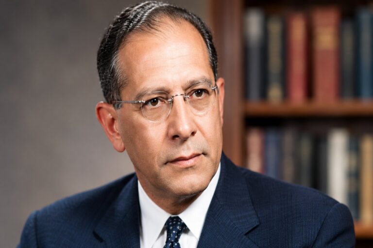 Albert Ezerzer in a formal portrait wearing a dark suit and glasses against a neutral background.